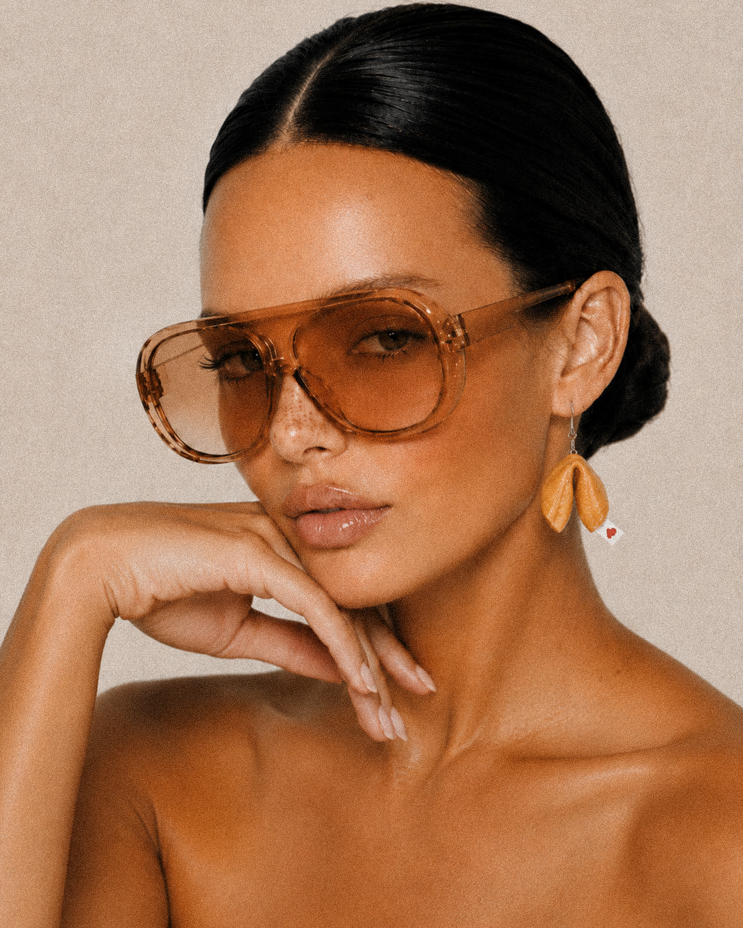 A close-up lifestyle shot of a woman with black hair in a bun sleek back wearing translucent amber brown tinted sunglasses and a fortune cookie earring resting her head on her hand.