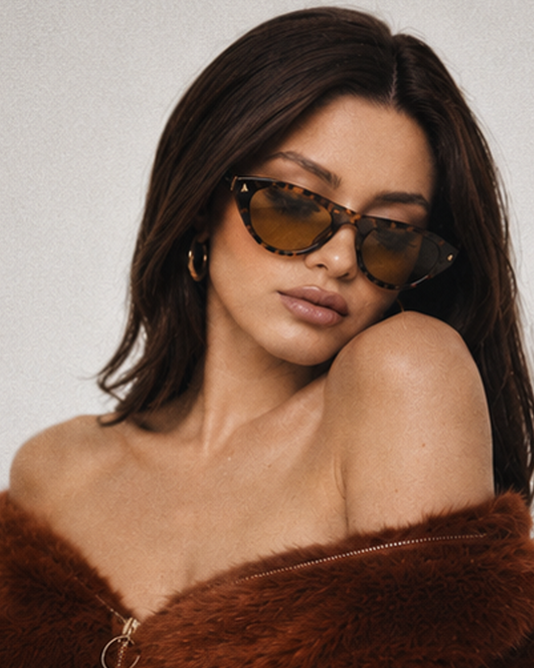 A woman with dark brown hair wearing cat-eye sunglasses with tortoiseshell frames and amber-tinted lenses, posed against a light gray background.