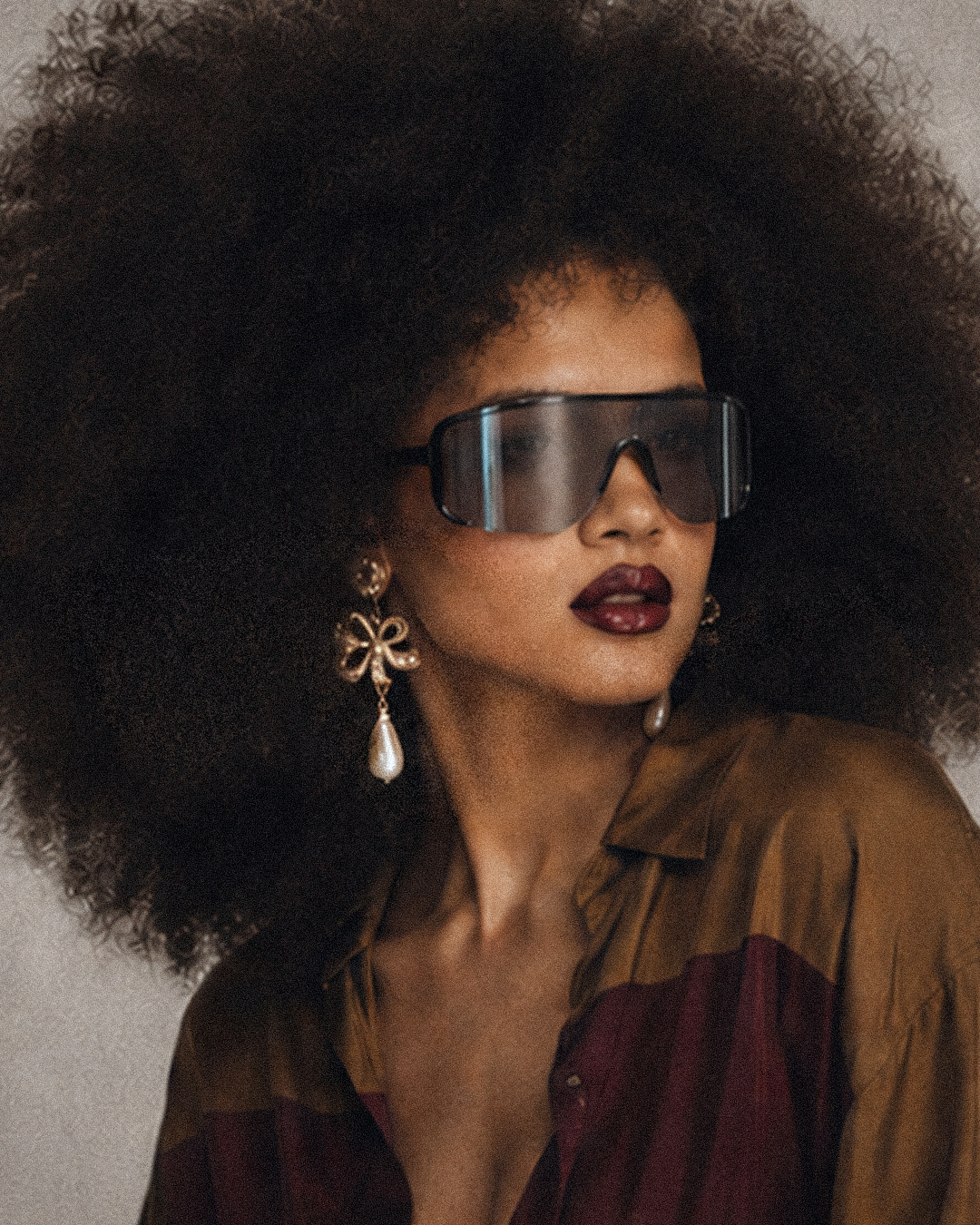 A close-up portrait of a model with a large dark afro hairstyle, wearing oversized dark-tinted shield sunglasses, dangling earrings, and a brown and burgundy satin shirt against a neutral light gray background.