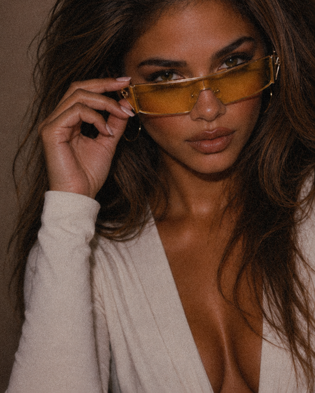 A close-up lifestyle shot of a woman with long brown hair wearing translucent amber-yellow tinted sunglasses and a cream-colored knit top, holding the sunglasses with her hand.