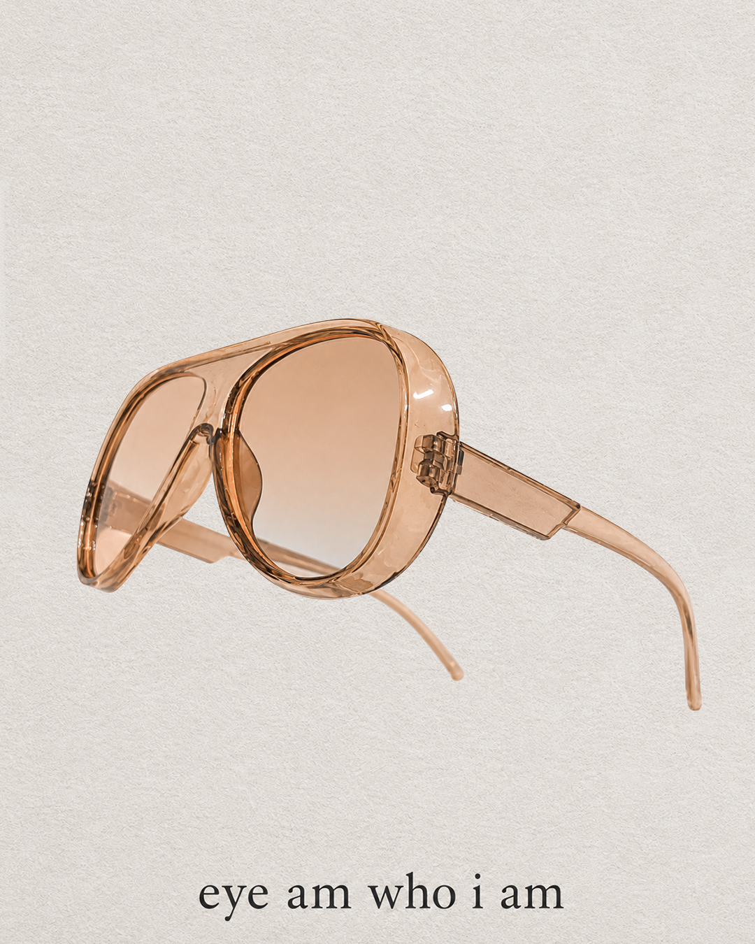 Oversized sunglasses with a translucent peach-pink frame and gradient tinted lenses, shown on a textured off-white background with the text 'eye am who i am' below.
