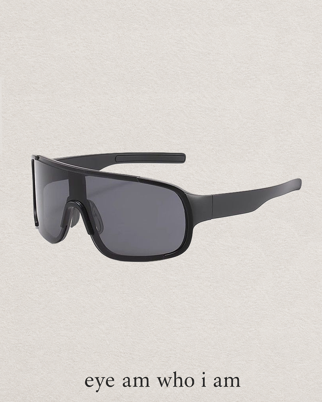 A pair of wraparound shield-style sunglasses with a matte black frame and dark gray lenses, shown on a light textured beige background with the text 'eye am who i am' below.